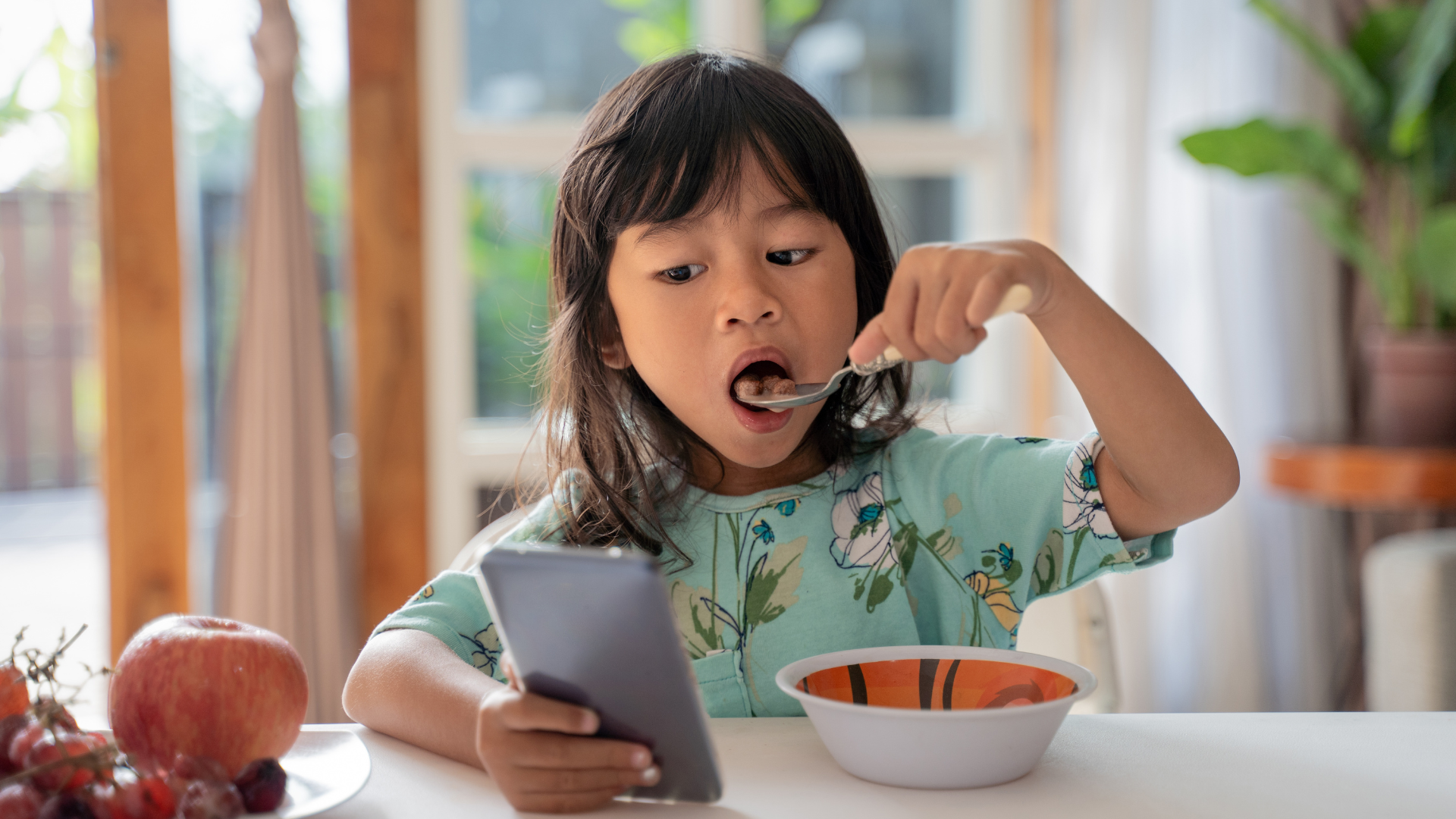 Young Child looking at phone in one hand while eating cereal with the other